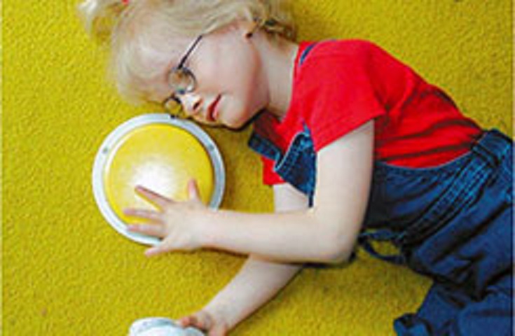 Ein Kind liegt auf einem Teppich und drückt mit der linken Hand auf einen großen Taster, während es in der linken Hand einen Talker hält