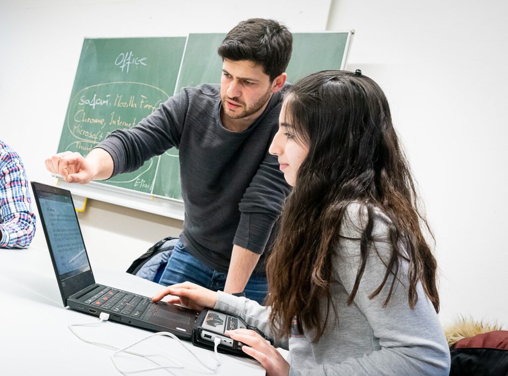 Die Schülerin Nariman Rashid sitzt vor ihrem Laptop. Ihr Beratungslehrer, Artjom Krichewskij (LFS) demonstriert Techniken im Umgang mit dem Laptop.