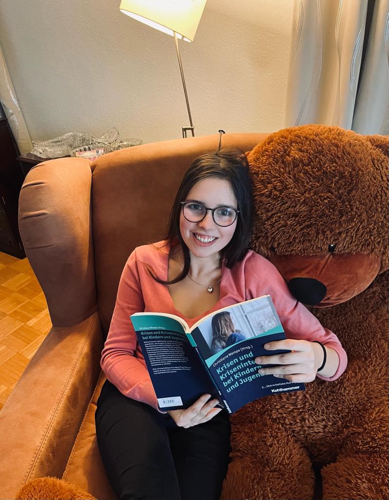 Julia sitzt in einem Sessel, neben sich einen Teddy ihrer Größe. In den Händen hält sie ein Fachbuch mit dem Titel "Krisen und Krisenintervention bei Kindern und Jugendlichen".