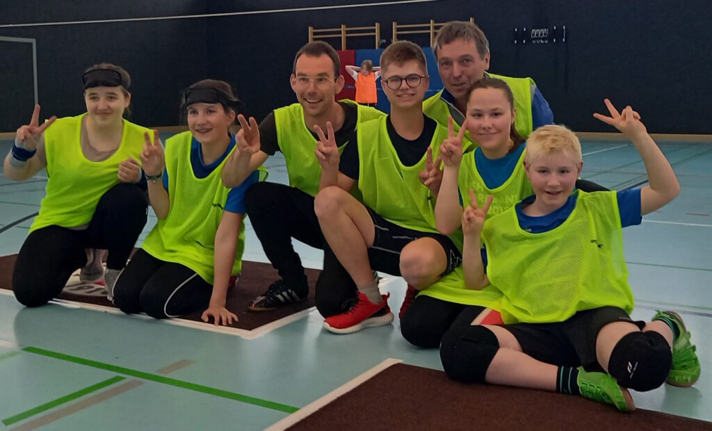 5 Schülerinnen und Schüler sowie zwei Lehrkräfte des LFS sitzen nebeneinander auf dem Hallenboden und schauen in die Kamera.