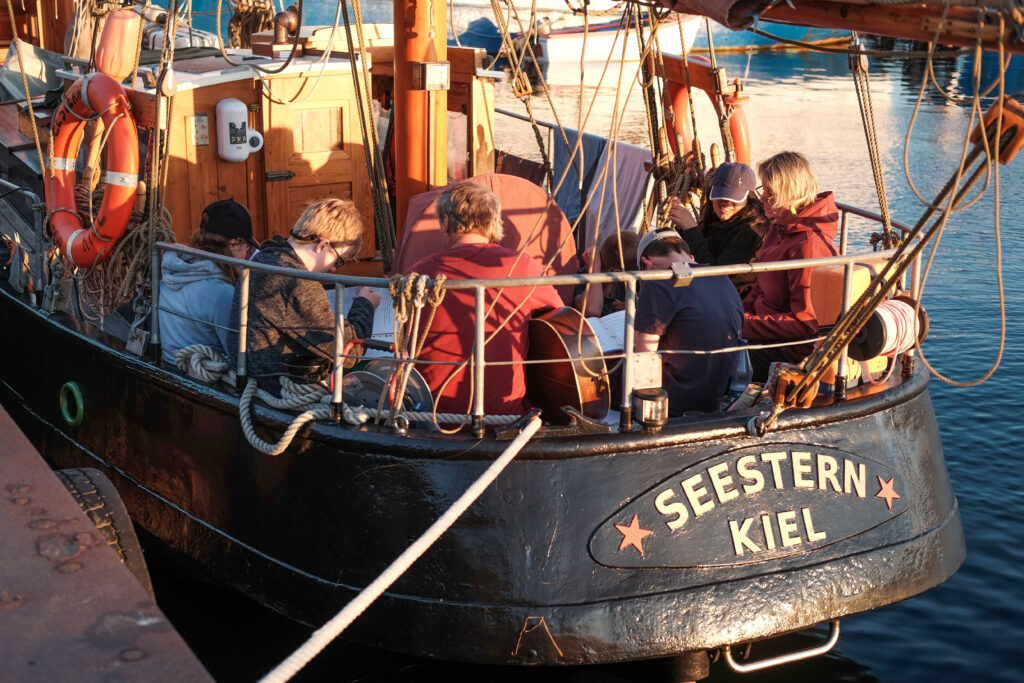 Das Segelschiff "Seestern Kiel" am Anleger. An Board befindet sich die Crew aus 5 Schülerinnen und Schülern des LFS sowie den kursdurchführenden Lehrkräften. Die Schülerinnen und Schüler haben vor sich einen Liedertext.