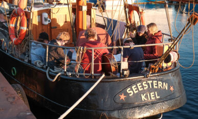 Das Segelschiff "Seestern Kiel" am Anleger. An Board befindet sich die Crew aus 5 Schülerinnen und Schülern des LFS sowie den kursdurchführenden Lehrkräften. Die Schülerinnen und Schüler haben vor sich einen Liedertext.