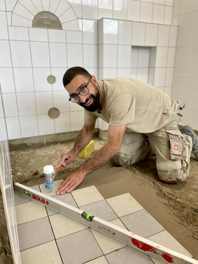 Masood kniet in der Bauhalle an seinem Arbeitsplatz. Er verlegt Bodenfliesen, vor sich eine Wasserwaage.