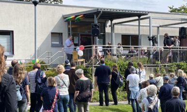 Frank Laemers, PH Heidelberg steht vor einem Rednerpult auf der Terrasse des Kurshauses. Weitere offizielle geladene Gäste sitzen auf Stühlen daneben. Davor stehen weitere Personen.