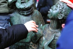 Kinderhände berühren eine bronzene Kinderstatue, die Teil einer Skulpturengruppe ist. Die Statue zeigt ein Kind mit nach oben gerichtetem Blick und geöffneten Händen, was ein Gefühl des Staunens vermittelt.