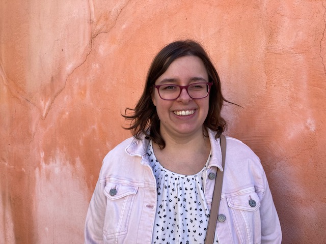 Sophie Gaden lächelt in die Kamera. Sie trägt eine hellrosa Jacke über einem weiß gemusterten Hemd und steht vor einer strukturierten orangefarbenen Wand.