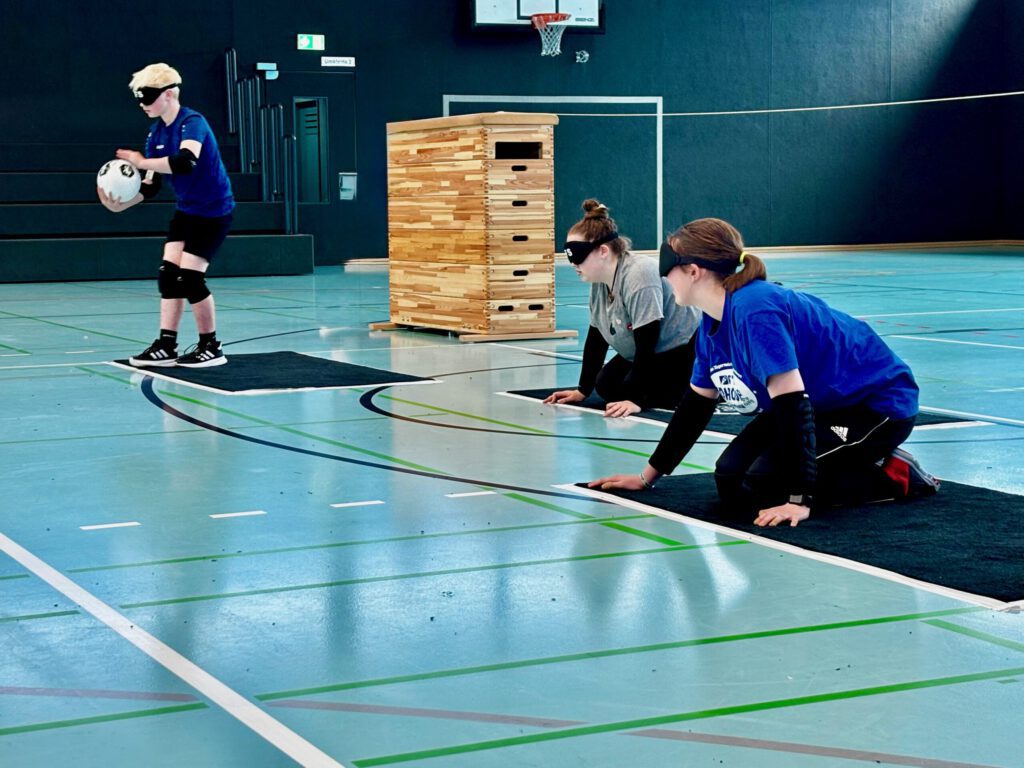 Drei Sportler mit Augenbinden, Knieschützern und Sportkleidung spielen in einer Turnhalle Goalball. Ein Spieler bereitet sich darauf vor, den Ball zu rollen, während zwei andere auf dem Boden knien und aufmerksam zuhören. Auf dem Bild sind zwei Spielerinnen und ein Spieler abgebildet. Alle befinden sich auf dünnen Fussmatten. Alle haben Augenbinden auf. Der Spieler ist links positioniert und hält einen Torball in den Händen. Er steht auf seiner Matte. Die Spielerinnen knien auf ihren Matten. Eine Spielerin befindet sich in der Mitte, die andere Spielerin befindet sich auf der rechten Seite.