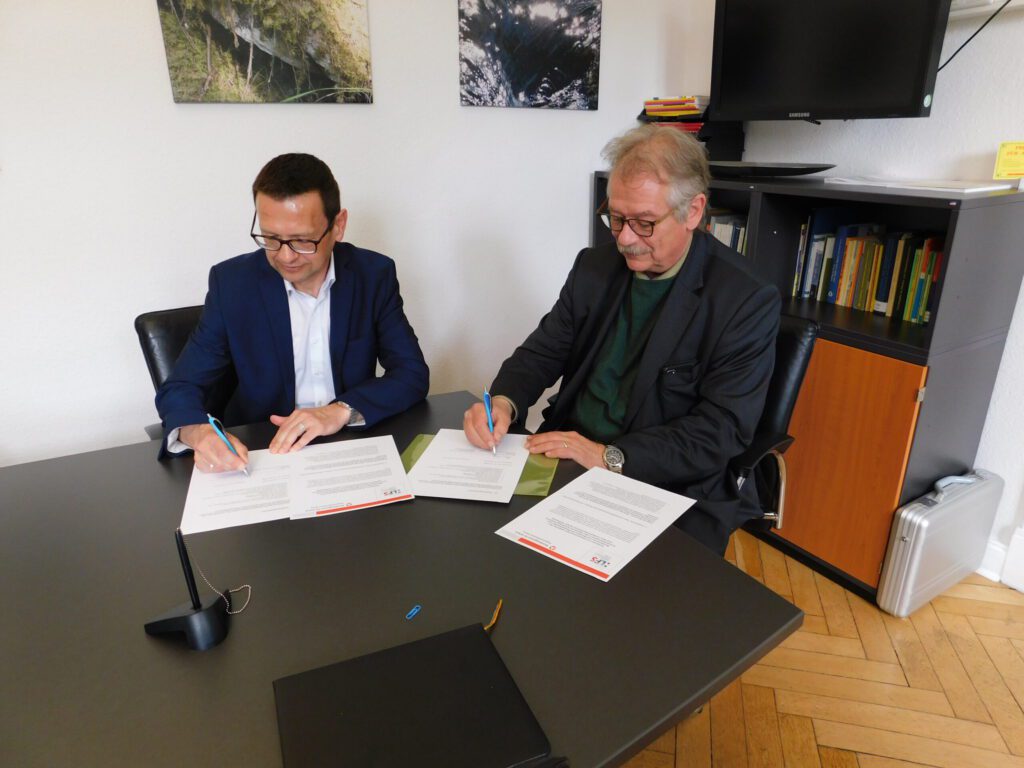 Zwei Männer in Geschäftskleidung sitzen an einem Tisch und unterschreiben Dokumente. Sie konzentrieren sich auf die vor ihnen liegenden Papiere. Im Hintergrund stehen ein Schrank mit Büchern und ein Fernseher, an der Wand hängen gerahmte Fotos. Zwei Männer in Geschäftskleidung sitzen an einem Tisch und unterschreiben ein Dokument.
