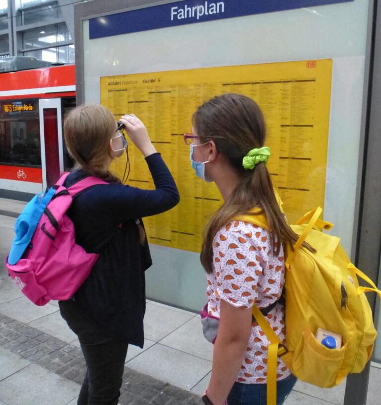 Zwei Schülerinnen am Bahnhof vor einem Fahrplan. Ein Mädchen benutzt ein Monokular, um den Fahrplan zu lesen.