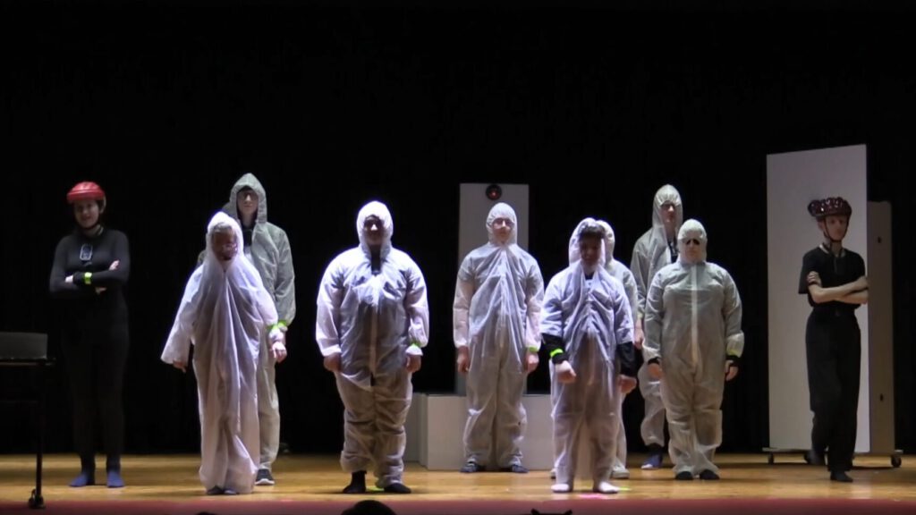 LFS-Theaterstück "Für dein vollkommenes Wohl" Eine Gruppe von Schülerinnen und Schülern in weißen Schutzanzügen steht auf einer Bühne, zwei weitere Personen in schwarzen Uniformen stehen an den Seiten. Der Hintergrund ist dunkel, und die Bühne ist mit Scheinwerfern beleuchtet.