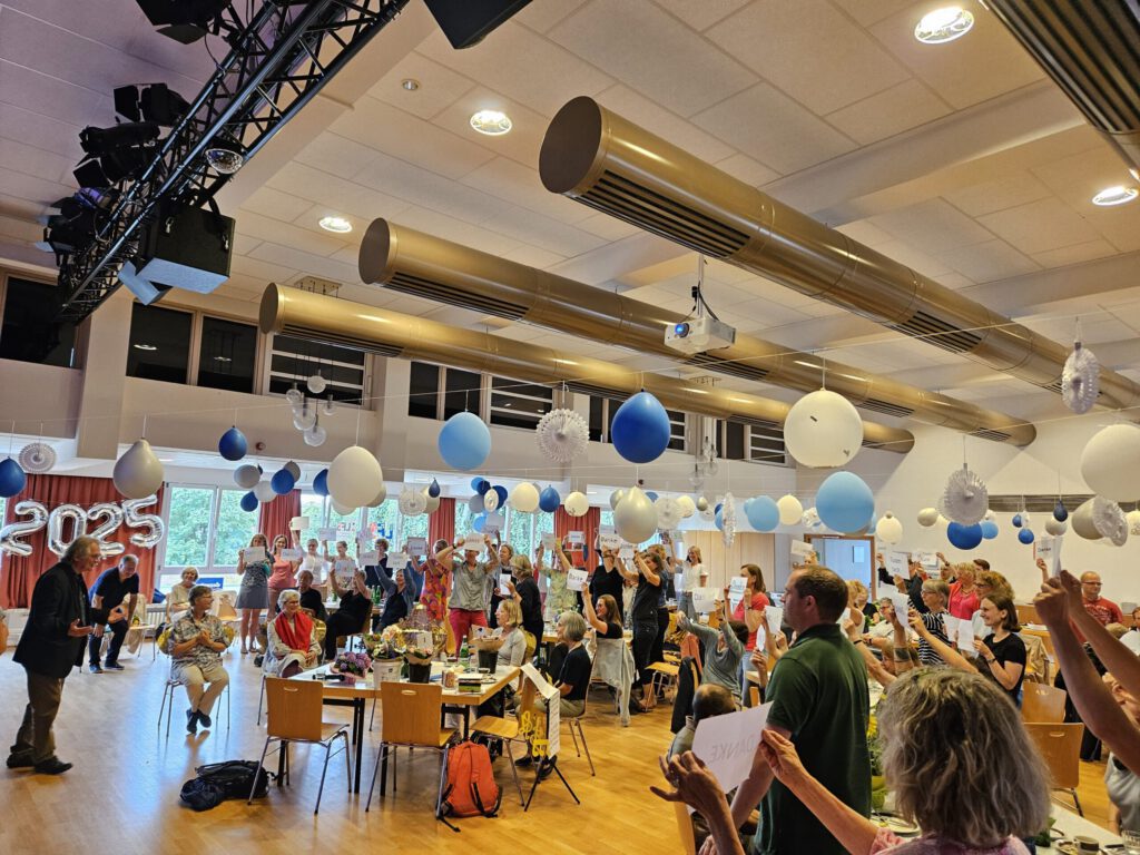 Das Kollegium sowie Ehemalige des LFS in der geschmückten Aula in Richtung Klaus Wißmann blickend, dabei ein Schild mit der Aufschrift "Danke" vor sich haltend.