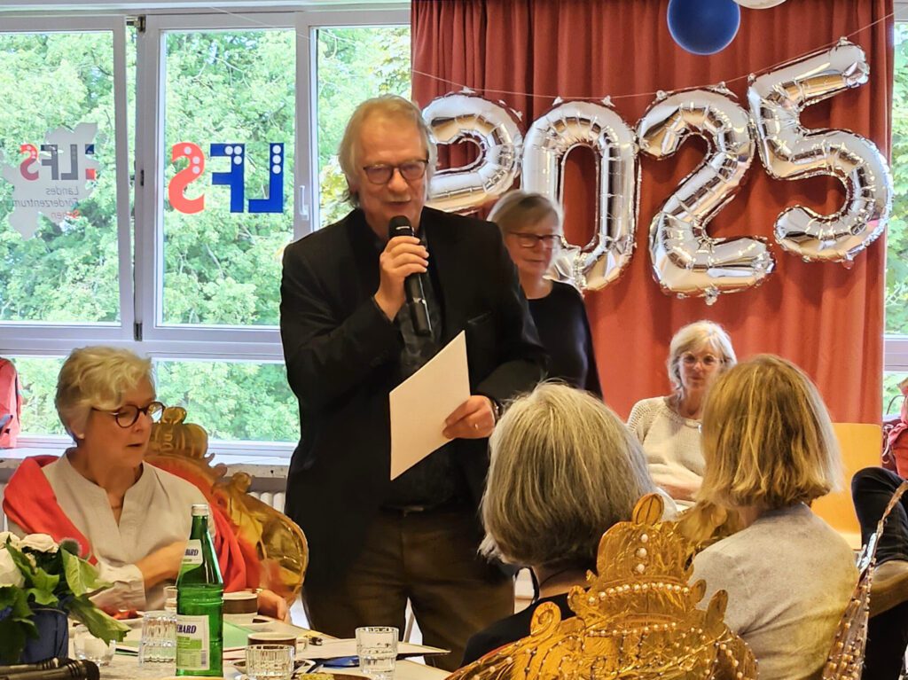 Klaus Wißmann steht in der Aula mit einem Mikrofon und einer Mappe in der Hand und spricht zum Kollegium. Die Kolleg*innen um ihn herum hören ihm aufmerksam zu.