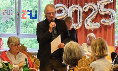 Klaus Wißmann steht in der Aula mit einem Mikrofon und einer Mappe in der Hand und spricht zum Kollegium. Die Kolleg*innen um ihn herum hören ihm aufmerksam zu.