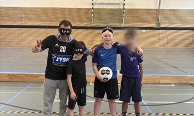 Auf dem Bild ist die Mannschaft des Landesförderzentrums Sehen zu sehen. Sie besteht aus drei Spielern und ihrem Trainer. Alle stehen auf einer dünnen Fußmatte mit der Aufschrift „MTV Heide von 1860“. Ein Spieler hält einen Torball in den Händen. Der Trainer und ein Spieler tragen Augenbinden mit der Aufschrift "LFS".