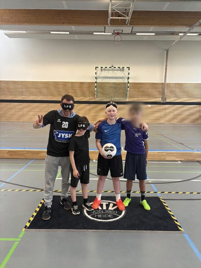 Auf dem Bild ist die Mannschaft des Landesförderzentrums Sehen zu sehen. Sie besteht aus drei Spielern und ihrem Trainer. Alle stehen auf einer dünnen Fußmatte mit der Aufschrift „MTV Heide von 1860“. Ein Spieler hält einen Torball in den Händen. Der Trainer und ein Spieler tragen Augenbinden mit der Aufschrift "LFS".