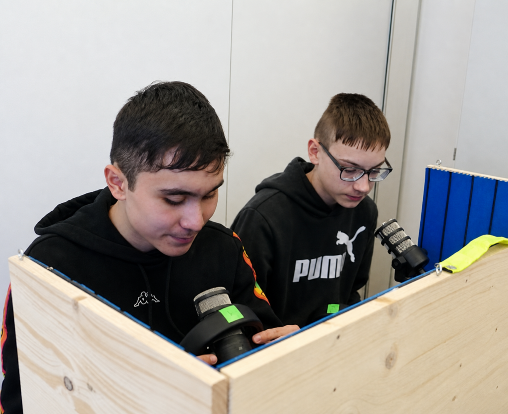 Zwei Schüler sitzen hinter einer Holzabtrennung und arbeiten mit Mikrofonen und technischen Geräten an einem Tisch.