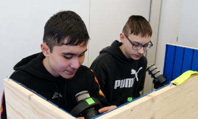 Zwei Schüler sitzen hinter einer Holzabtrennung und arbeiten mit Mikrofonen und technischen Geräten an einem Tisch.