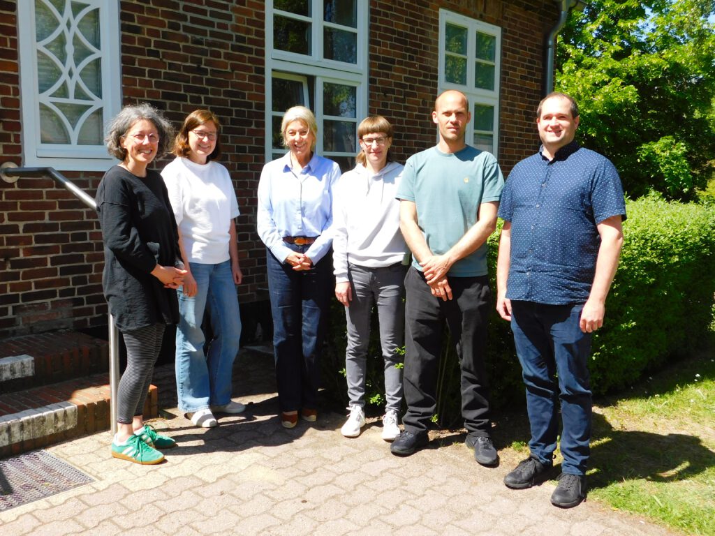 Auf dem Bild sind von links nach rechts: Nicole Hinsch-Tönnies, Katharina Bossert De Paz, Michaela Pries, Lou B., Moritz Magnussen, Björn Albrecht Im Hintergrund sieht man einen Ausschnitt eines roten Backsteingebäudes, das Verwaltungsbebäude des LFS sowie grüne Büsche.