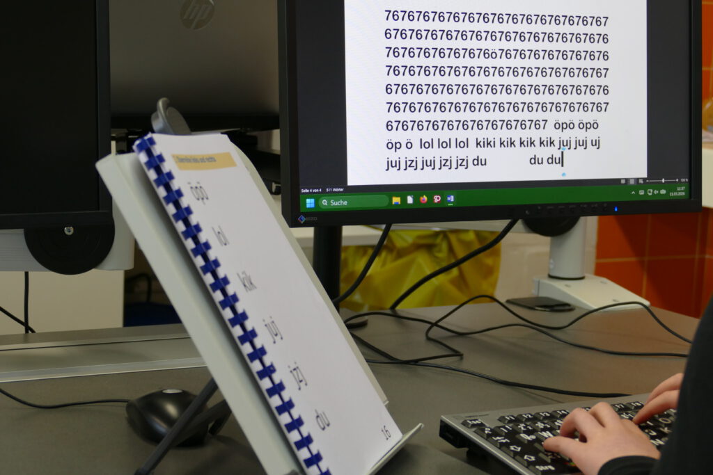 Schüler sitzt an einer Tastatur und überträgt einen Text von einem Ausdruck auf den PC. Auf dem Bildschirm ist die Übertragung in vergrößerter Schrift zu sehen.