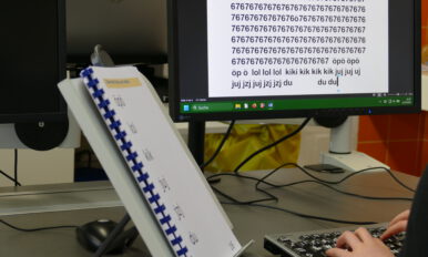 Schüler sitzt an einer Tastatur und überträgt einen Text von einem Ausdruck auf den PC. Auf dem Bildschirm ist die Übertragung in vergrößerter Schrift zu sehen.