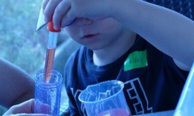Ein Kind hält eine Pipette über ein Glas und untersucht orange Flüssigkeit bei einem Experiment.