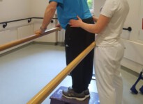Hier trainiere ich mit einem Patienten die Muskulatur des Standbeines als Vorübung zum Treppensteigen. Um die aufrechte Körperposition entsprechend korrigieren zu können, lege ich meine Hände an das Becken des Patienten.