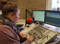 Die Musik wird über das Mischpult gesteuert und anschließend im Radio ausgestrahlt. Seitlicher Blick auf die Schülerin, die vor einem Mischpult und Mikrofon sitzt. Im Hintergrund sind zwei Bildschirme zu sehen.