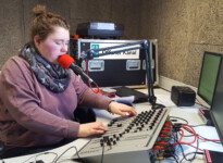 Auf diesem Bild spreche ich gerade Kinonachrichten oder Ansagen für Musiktitel in das Mikrofon. Schülerin sitzt vor dem Mischpult und Mikrofon und hält die Hände an den Reglern