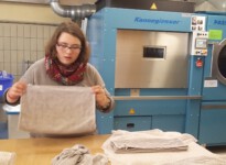 Die Wäschestücke müssen immer nach dem gleichen Schema zusammengelegt werden. Anna hält ein Handtuch in beiden Händen und orientiert sich dabei an den umgenähten Kanten. Im Hintergrund sieht man große Industriewaschmaschinen. junge Frau vor einem Trockner, die Handtücher faltet