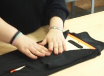 Anna legt ein T-Shirt zusammen. Gute Kontraste und ein Legebrett erleichtern ihr diese Tätigkeit. Auch die T-Shirts müssen immer auf die gleiche Weise zusammengelegt werden, damit sie anschließend exakt gestapelt und für den Kunden verpackt werden können. junge Frau faltet ein Hemd zusammen
