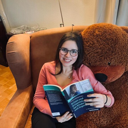 Julia sitzt in einem Sessel, neben sich einen Teddy ihrer Größe. In den Händen hält sie ein Fachbuch mit dem Titel "Krisen und Krisenintervention bei Kindern und Jugendlichen".