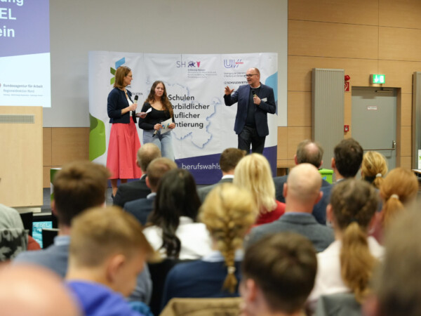 Die Veranstalter*innen der Auszeichnung, Frau Dose und Herr Gaede sowie eine Schülerin, die ebenfalls die Veranstaltung moderiert, stehen auf einer Bühne vor einem Publikum und sprechen. Im Hintergrund ist ein Banner mit der Aufschrift „Schulen mit vorbildlicher beruflicher Orientierung“ zu sehen, das Logos von Institutionen wie dem Ministerium für Bildung Schleswig-Holstein, der Bundesagentur für Arbeit sowie der Vereinigung der Unternehmensverbände in Hamburg und Schleswig-Holstein enthält. I