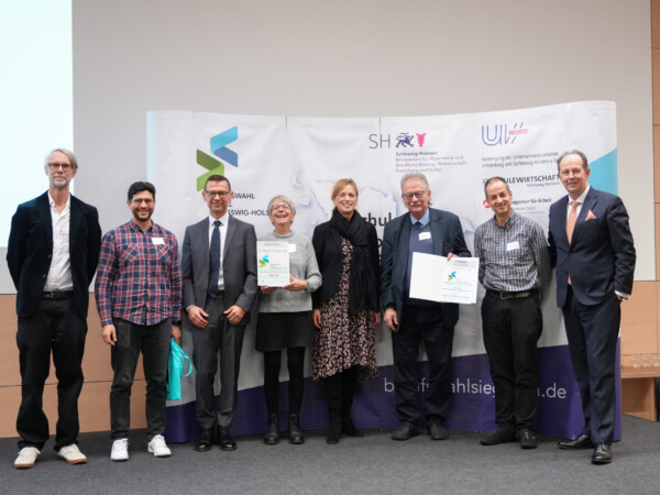 Eine Gruppe von acht Personen, darunter Herr Radeiske (Kreisfachberaterinnen und Kreisfachberater für Berufliche Orientierung), Herr (BA, Regionaldirektion Nord), Frau Prien (Bildungsministerin), Herr Fröhlich (UV Nord) sowie vom LFS Herr Wißmann, Herr Hilgers, Frau Brodersen und Herr Krichewskij, steht vor einem Banner mit der Aufschrift „Berufswahlsiegel Schleswig-Holstein“ und weiteren Logos, darunter das des Ministeriums für Bildung Schleswig-Holstein. Frau Brodersen hält das Berufswahlsiegel-Schild mit der Aufschrift "2. Rezertifizierung" und Herr Wißmann die dazugehörige Urkunde. Alle lächeln in die Kamera und stehen in festlicher Kleidung auf einer Bühne.