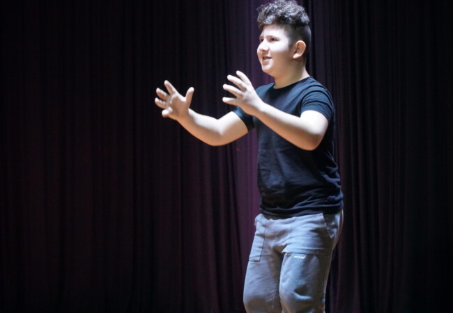 Schüler in Aktion während eines Theaterauftritts. Ein junger Schauspieler in schwarzem T-Shirt und grauen Hosen, der auf einer Bühne mit ausgebreiteten Armen steht und eine Darbietung macht.