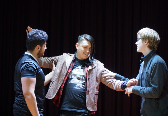 Drei Personen auf der Bühne in Interaktion. Drei Schülerinnen und Schüler einer Theatergruppe führen eine Szene auf. Eine Person in der Mitte trägt eine gemusterte Jacke und ein kariertes Hemd und wird von den anderen beiden Personen an den Armen festgehalten. Die Szene spielt vor einem dunklen Vorhang und wirkt intensiv und emotional.