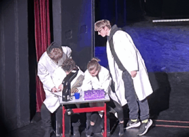 Vier Personen in weißen Laborkitteln versammeln sich um einen Tisch auf einer Bühne. Drei untersuchen etwas unter dem Mikroskop, eine Person steht daneben.