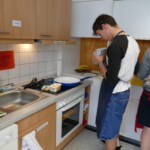 Eine Person, die ein schwarz-weißes Hemd und Jeans-Shorts trägt, steht an einer Küchentheke und bereitet Essen zu, wobei eine weitere Person teilweise neben ihr zu sehen ist. Die Küche ist modern mit Holzschränken und einem Ofen.