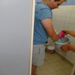 Eine Person in einem blauen Hemd und grauen Shorts steht in einem gekachelten Raum und nimmt über einem Mülleimer rosa Gummihandschuhe aus den Händen einer anderen Person entgegen. Die Person trägt leuchtend gelb-grüne Schuhe.