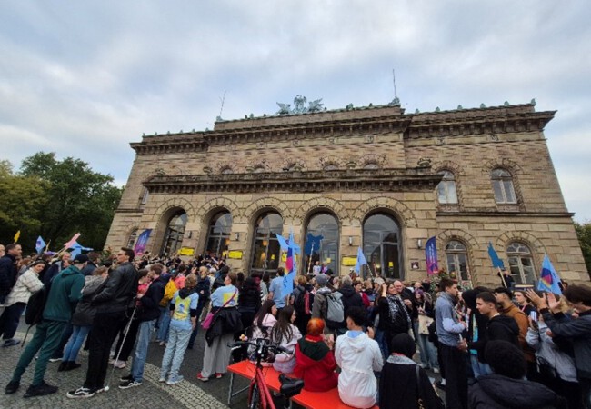 Viele Menschen stehen mit bunten Fahnen und Transparenten vor einem historischen Theatergebäude. Die Atmosphäre ist lebendig.