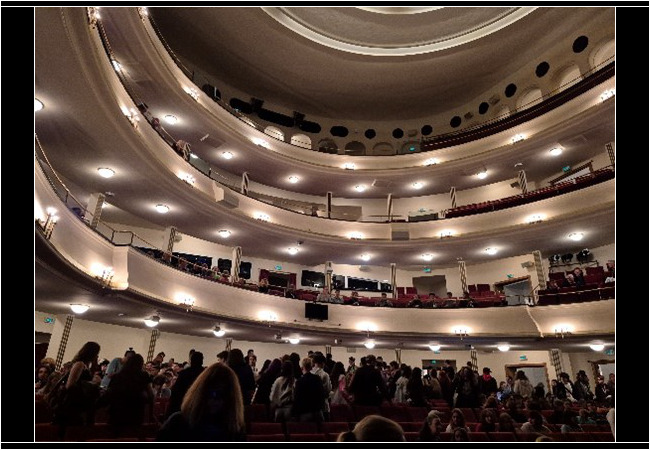 Blick ins Innere eines großen Theatersaals mit drei Rängen. Der Zuschauerraum ist gut gefüllt, viele Menschen stehen oder suchen ihren Platz. Die Beleuchtung ist warm, die Architektur elegant geschwungen.