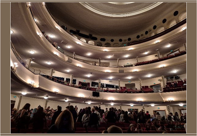 Blick ins Innere eines großen Theatersaals mit drei Rängen. Der Zuschauerraum ist gut gefüllt, viele Menschen stehen oder suchen ihren Platz. Die Beleuchtung ist warm, die Architektur elegant geschwungen.