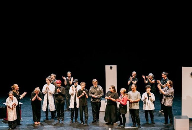 Die LFS Theatergruppe mit etwa fünfzehn Personen steht auf einer Bühne und applaudiert in Richtung der Regisseurin, Martina Müller-Korn (LFS). Einige tragen helle Kittel oder schwarze Kleidung. Im Hintergrund ist die Bühne schwarz gehalten.