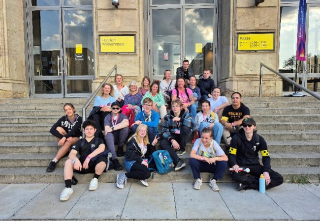 Die LFS-Theatergruppe sitzt entspannt auf einer Treppe vor dem Theatergebäude. Sie schauen in die Kamera und lächeln, im Hintergrund sind gelbe Schilder mit der Aufschrift „Herzlich willkommen!“ zu sehen.