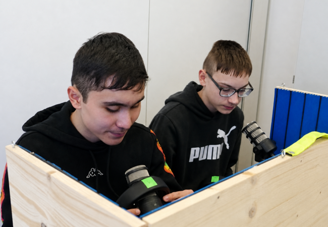 Zwei Schüler sitzen hinter einer Holzabtrennung und arbeiten mit Mikrofonen und technischen Geräten an einem Tisch.