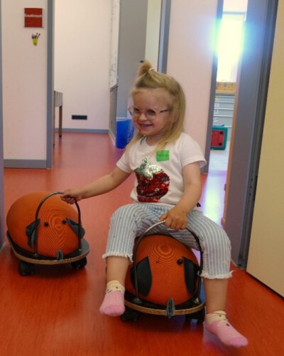 Ein lächelndes Kind sitzt auf einem Rutschauto und zieht im Kurshausflur ein weiteres Rutschauto hinter sich her