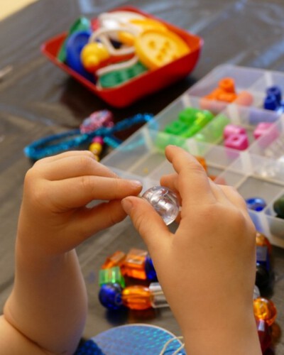 Kinderhände ziehen einen Faden durch eine durchsichtige große Perle neben einer Sortierbox mit weiteren bunten Perlen