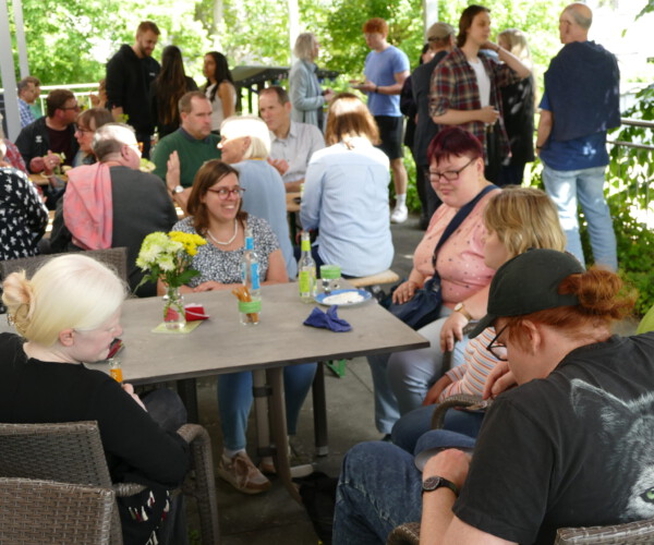 Die Terrasse hat sich mit Menschen gefüllt.
Eine Markise schützt vor der Sonne.